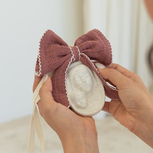 Bendición de cuna personalizada con moño color borgoña sostenida en manos, detalle de medalla de la Virgen con el Niño