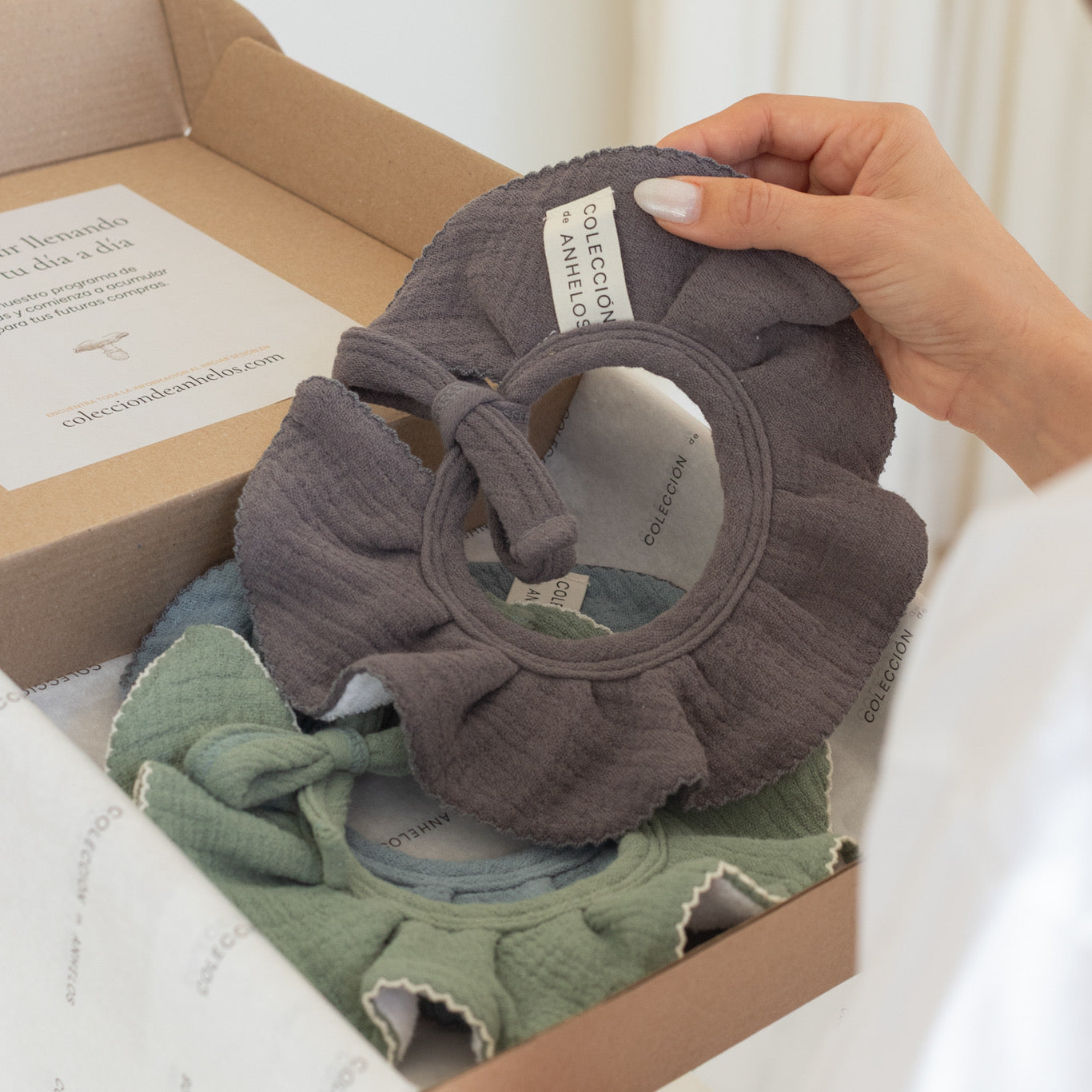 Cuello babero para bebé en color gris siendo sacado de una caja por una mano