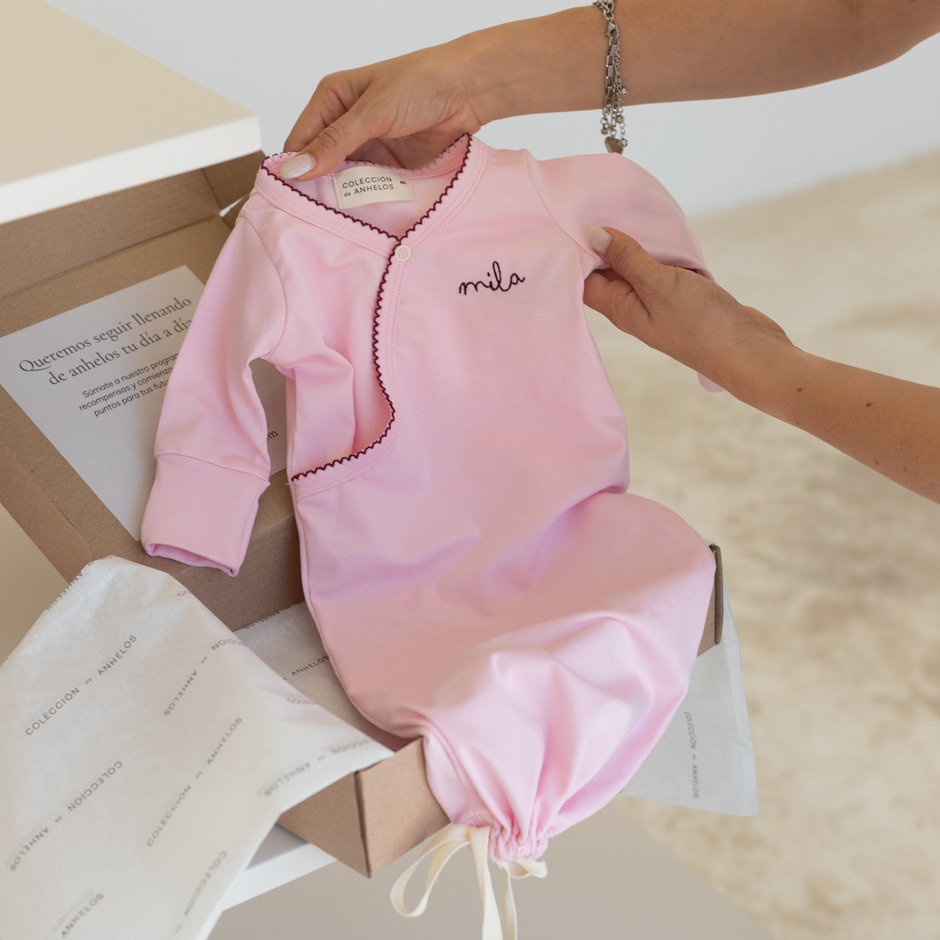 Pijama costalito para bebé en color rosa con bordado borgoña siendo sacada de una caja por una mamá